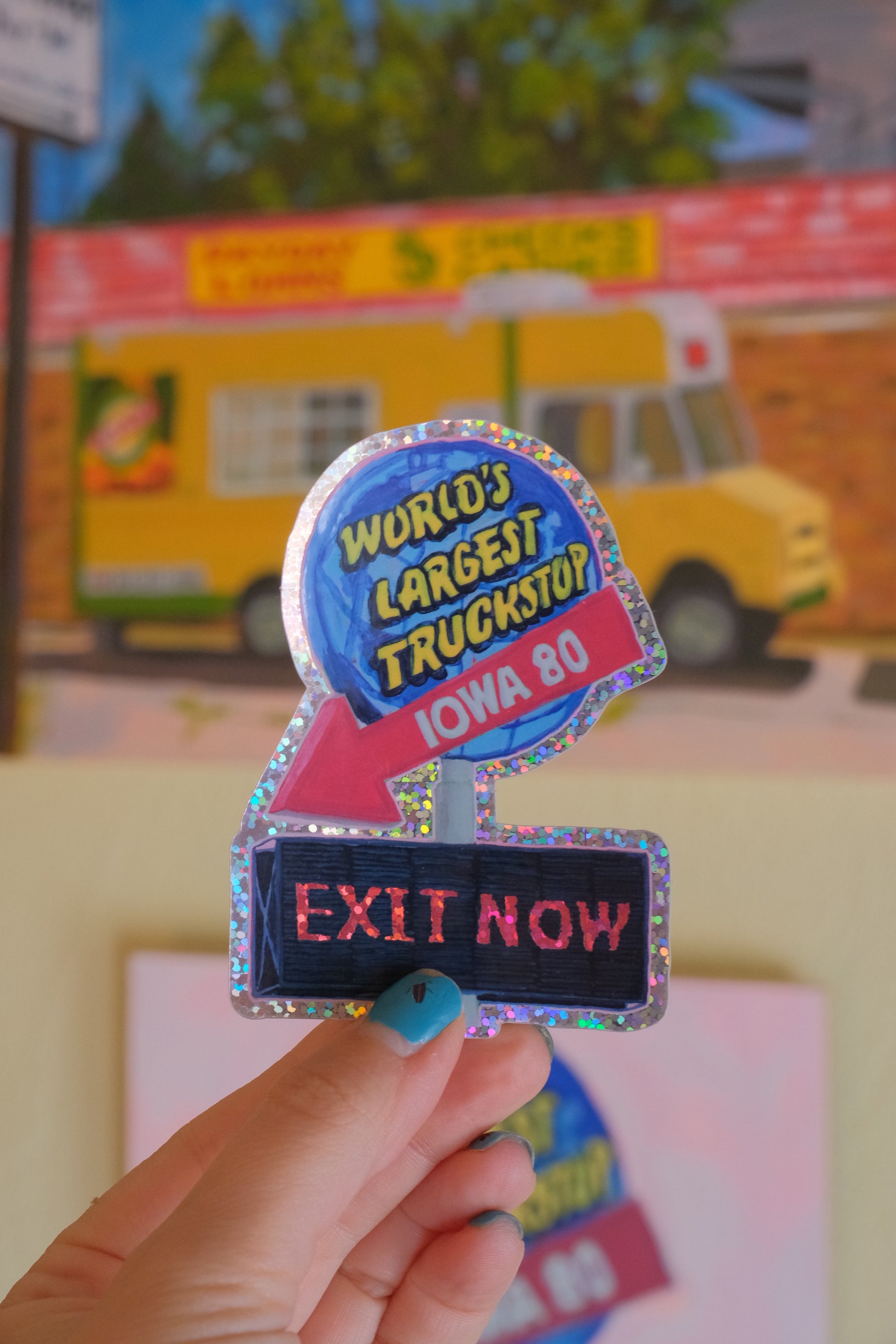 A hand holding a novelty trucker-style sticker or patch with a holographic border, featuring a blue circular badge that reads 'World's Largest Truck Stop IOWA 80' and a pink arrow pointing to a dark blue vertical sign that says 'EXIT NOW'.
